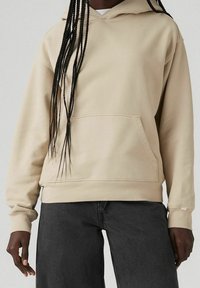 Felpa beige con cappuccio, tasca frontale, polsini a costine e vestibilità morbida, abbinata a jeans larghi grigio scuro.