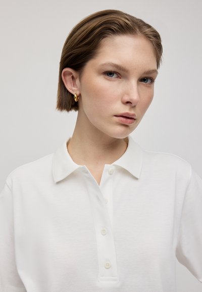 Polo shirt blanc en tissu texturé, avec un col classique et une patte de boutonnage à trois boutons, porté par une personne aux cheveux courts. Boucles d'oreilles en or en forme de créole.