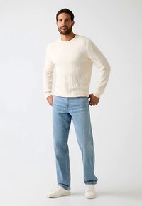 Pull crème en maille torsadée à col rond, associé à un jean droit bleu clair et des baskets blanches. Sur un fond neutre.