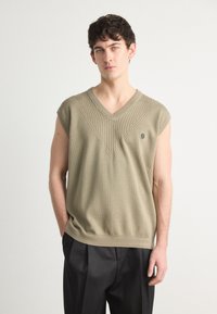 Pulôver sem mangas em verde oliva, com um decote em V e nervuras texturizadas no peito, apresentando um logótipo subtil no lado esquerdo.