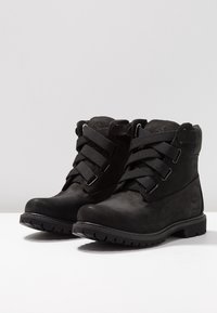 Black ankle boots made of leather with textile accents, featuring a lace-up design, thick rubber sole, and logo detailing on the side.