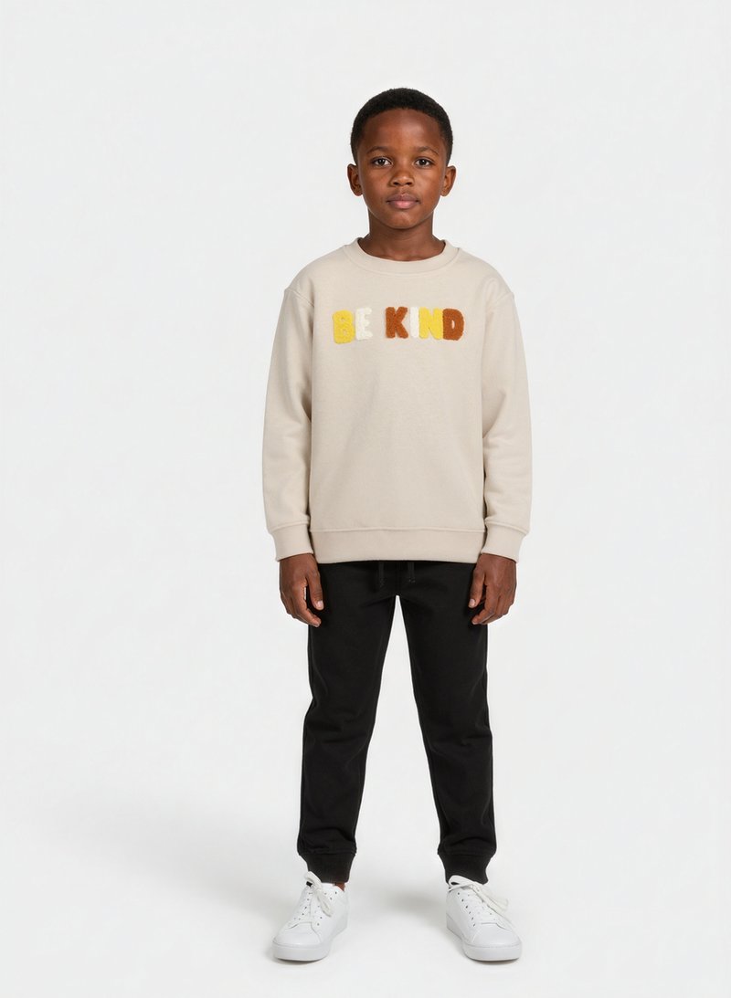Beige mikina s nápisem "BUĎ DOBRÝ" z plyšových písmen, v kombinaci s černými tepláky a bílými teniskami. Měkká textura, volný střih, pohodlný design.