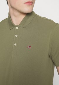 Oliven grønn polo skjorte laget av teksturert stoff, med klassisk krage og tre knapper, samt et lite brodert Norge-flagg logo.