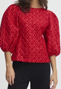 Haut rouge à manches bouffantes avec un motif floral texturé, un col rond et un design en tissu superposé. Associé à un pantalon noir à rayures.