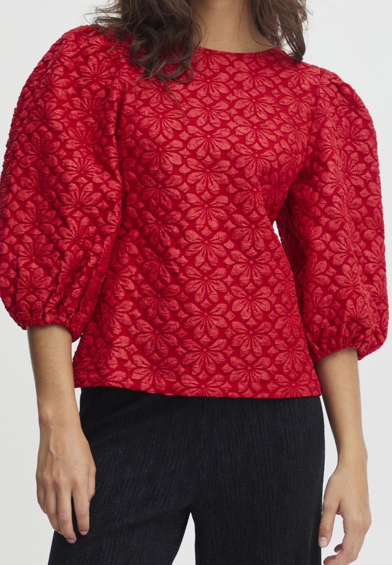 Haut rouge à manches bouffantes avec un motif floral texturé, un col rond et un design en tissu superposé. Associé à un pantalon noir à rayures.