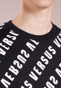 Felpa nera con collo a giro e audace testo diagonale bianco che legge "VERSUS," con una texture liscia e opaca.