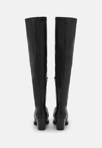 Botas de cuero negras hasta la rodilla con una textura suave, tacones altos de bloque y cremalleras en los laterales, con costuras verticales en la parte trasera.