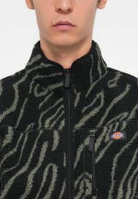 Black and olive green fleece jacket with a zipper, featuring a textured zebra pattern and a chest pocket with a logo.