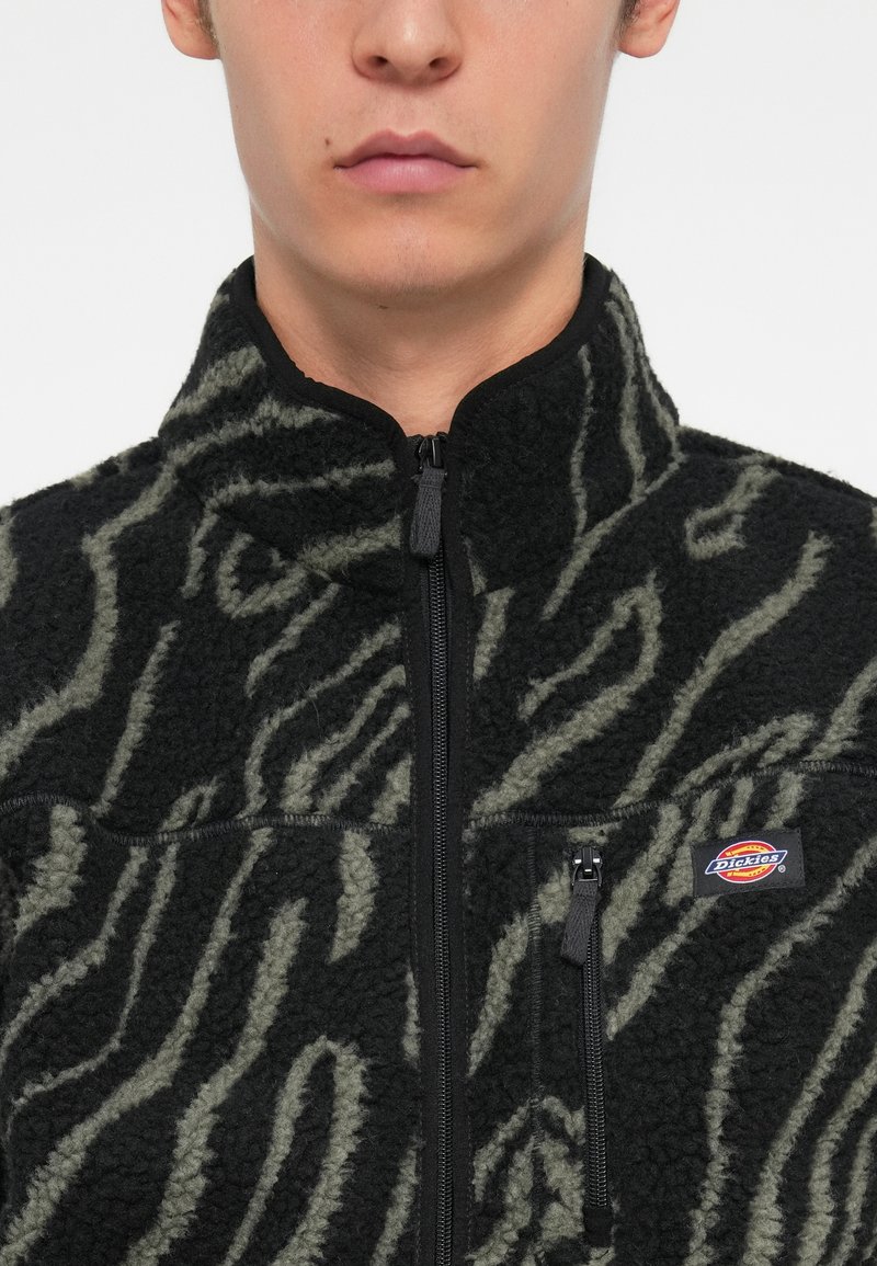 Sort og olivengrøn fleecejakke med lynlås, udstyret med et tekstureret zebra-mønster og en brystlomme med et logo.