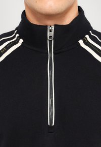 Svart zip-up jakke med høy krage. Har hvite aksentstriper på skuldrene og teksturert stoff. Merkbar metallglidelås.