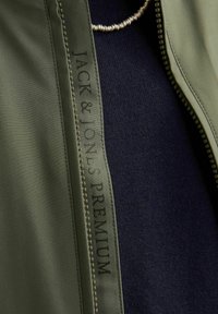 Olívazöld dzseki cipzárral és "JACK & JONES PREMIUM" felirattal ellátott címkével; belső oldala textúrázott sötétkék anyagból készült.