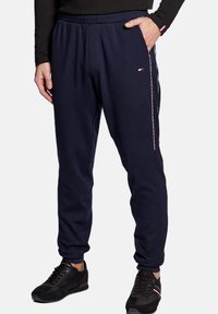 Pantalones de chándal azul marino con una textura suave, cinturilla elástica y detalles de franjas laterales en rojo y blanco. Cuenta con bolsillos frontales y un corte ajustado.
