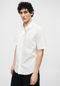 Νέος άνδρας με σγουρά μαλλιά, που φοράει λευκό πουκάμισο με κοντά μανίκια και στρογγυλό λογότυπο στο στήθος, συνδυασμένο με μαύρο παντελόνι.