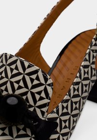 Chaussure à bride arrière noire et crème avec un motif géométrique, bout arrondi et nœud noir en accent. Tige en cuir et semelle intérieure beige.