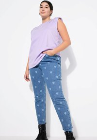 Lavendelfarbenes ärmelloses Oberteil mit besetzten Schultern, kombiniert mit hellblauen Jeans mit Herzmustern und schwarzen Knöchelstiefeln.