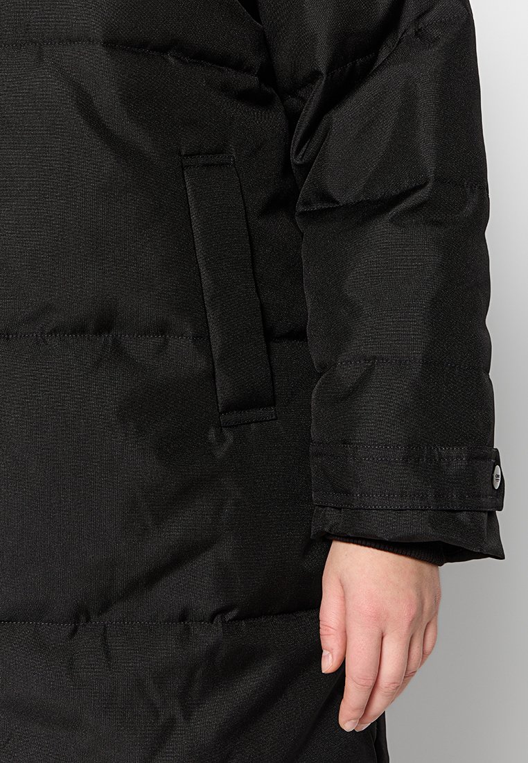 Détail de la manche et de la poche d'une veste noire matelassée, avec la main droite d'une personne visible sur fond blanc.