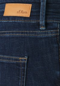 Tummansiniset farkut, joissa on ruskea s.Oliver-tarra, erottuva ommel ja sivutaskuyksityiskohta. Pehmeä materiaali ja klassinen malli.