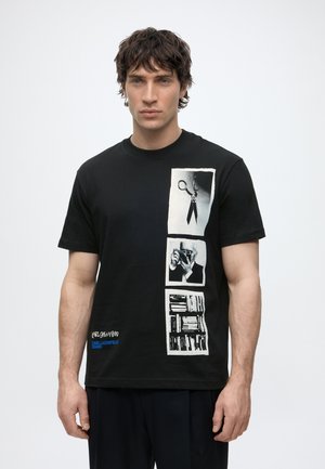 Mann trägt schwarzes T-Shirt mit vertikaler Collage aus Scheren, einer Person, die eine Kamera hält, und Büchern vorne, steht vor schlichtem Hintergrund.