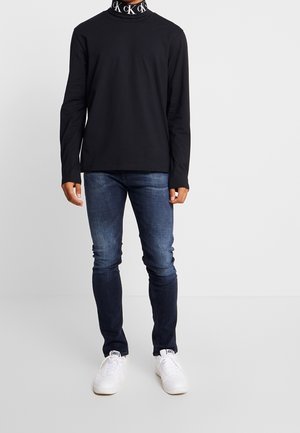 Black long-sleeve t-shirt with high collar featuring white logo detail, paired with dark blue fitted jeans and white sneakers.