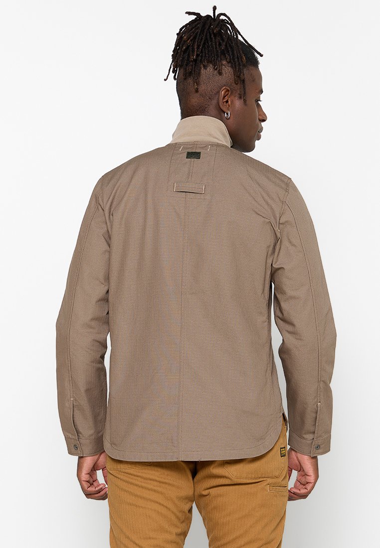 Chaqueta marrón hecha de un tejido texturizado, con un cuello medio, costuras verticales y una pequeña etiqueta con el logo en el cuello. Manga larga con puños de botón.