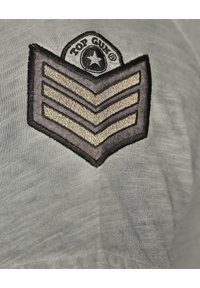 Grått tyg med en strukturerad, broderad lapptest med ett chevronmönster i beige och svart, märkt "Top Gun" med en stjärna.