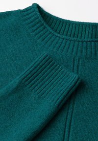 Teal stickad tröja med ribbad krage och muddar. Slät textur med subtil glans. Har ett stickat mönster längs ärmarna och kroppen.