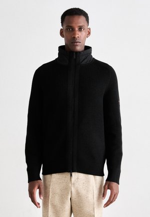 Schwarze Rippstrick-Zip-Jacke mit hohem Kragen; strukturiertem Stoff; figurbetonter Schnitt; getragen über hellen, strukturierten Hosen.