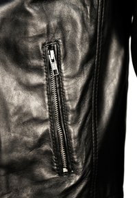 Sort læderjakke med en synlig sidelomme med sølvfarvet lynlås, glat tekstur og diskrete syninger langs sømmen.