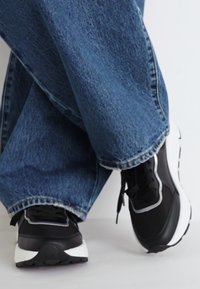 Svarte atletisk sko med hvite detaljer, laget av glatt materiale og med polstret tunge, kombinert med løse blå denimjeans.