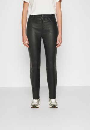 Personne portant un pantalon skinny noir en similicuir, un t-shirt blanc et des baskets blanches, debout devant un fond uni.