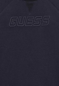 Sudadera azul marino con un logo texturizado "GUESS" en el pecho, hecha de una tela suave y lisa, con cuello redondo y mangas raglán.