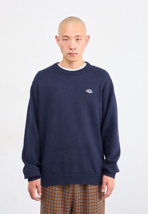 Man with shaved head wearing navy blue Dickies sweater and brown checkered pants, standing against white background.