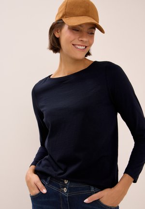 Femme portant une casquette marron, une chemise à manches longues bleu marine et un jean bleu, les mains dans les poches, regardant vers le bas et souriant.