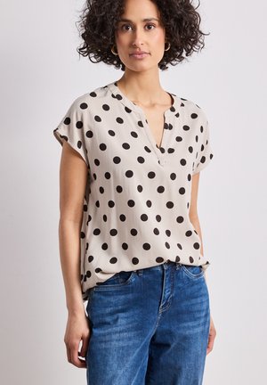 Femme aux cheveux bouclés portant un chemisier beige à manches courtes avec des pois noirs et un jean bleu, debout devant un fond neutre.