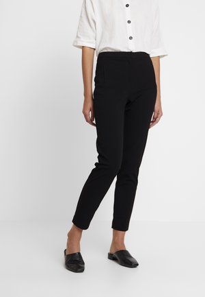 Person wearing black slim-fit ankle pants, white buttoned shirt with rolled sleeves, and black woven slip-on shoes, standing against plain background.