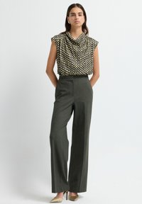 Femme portant un haut sans manches à motifs noir, blanc et jaune, un pantalon large gris, des boucles d'oreilles en or et des chaussures à talons beiges.