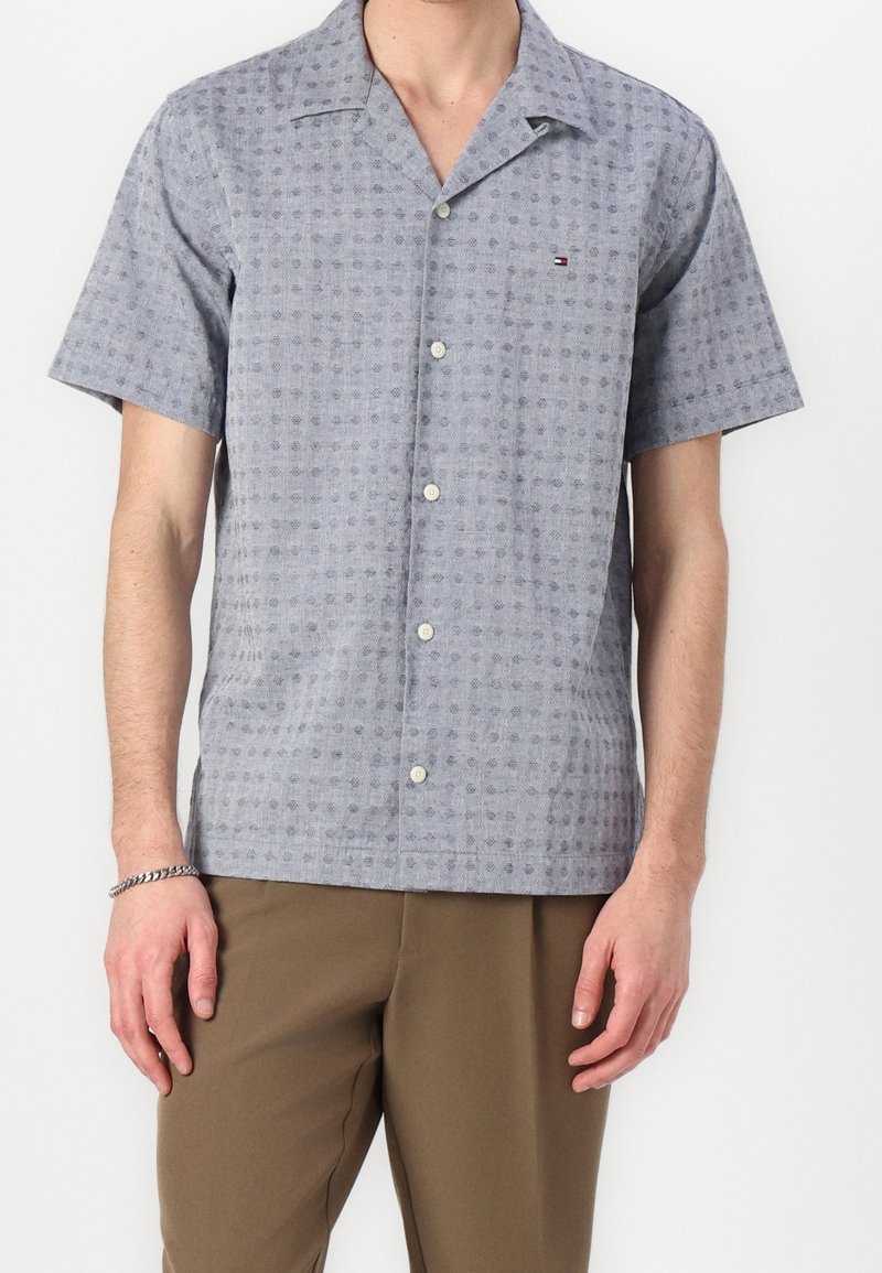 Homme portant une chemise à manches courtes bleu clair à boutons avec un motif géométrique subtil et un pantalon marron.