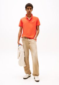 Oransje polo-skjorte med krage, beige vide jeans og hvite joggesko. En lysegrå hettegenser er drapert over armen.