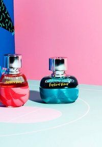 Två parfymflaskor: en röd och en turkos, båda med rundad form, metalliska silverlock och etiketter med texten "Festival Vibes".