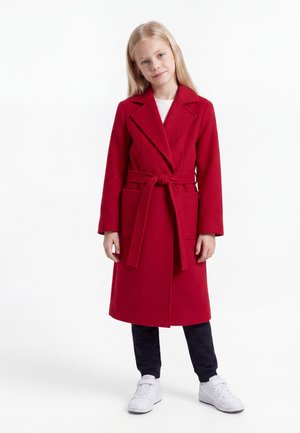 Young girl with long blonde hair wearing a red belted coat, black pants, and white sneakers standing against a white background.