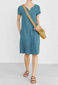 Vestido de lino verde azulado con mangas cortas, escote en V, bolsillos laterales y un corte relajado. Combinado con sandalias marrones de tiras y un bolso bandolera en color tierra.
