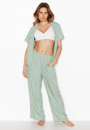 Vrouw draagt lichtgroene en witte geruite pyjamabroek en een open kort mouw hemd met een witte beha, staat blootsvoets tegen een witte achtergrond.