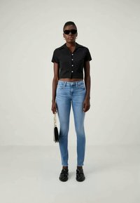 Camisa negra corta abotonada, jeans ajustados azul claros, zapatos negros, sosteniendo un pequeño bolso negro con cadena. La modelo lleva gafas de sol.