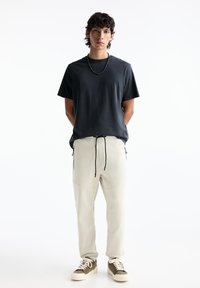 Dunkelgraues T-Shirt mit kurzen Ärmeln, kombiniert mit hellbeigen, schmal zulaufenden Sweatpants, die über eine verstellbare Kordel und seitliche Reißverschlusstaschen verfügen. Braune Sneaker.