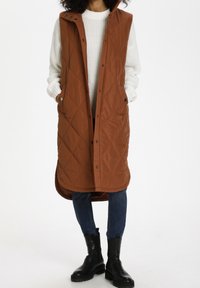 Brązowa pikowana długa kamizelka z kapturem, z przodu na zatrzaski i kieszeniami po bokach, noszona na białym swetrze i niebieskich dżinsach, zestawiona z czarnymi botkami.