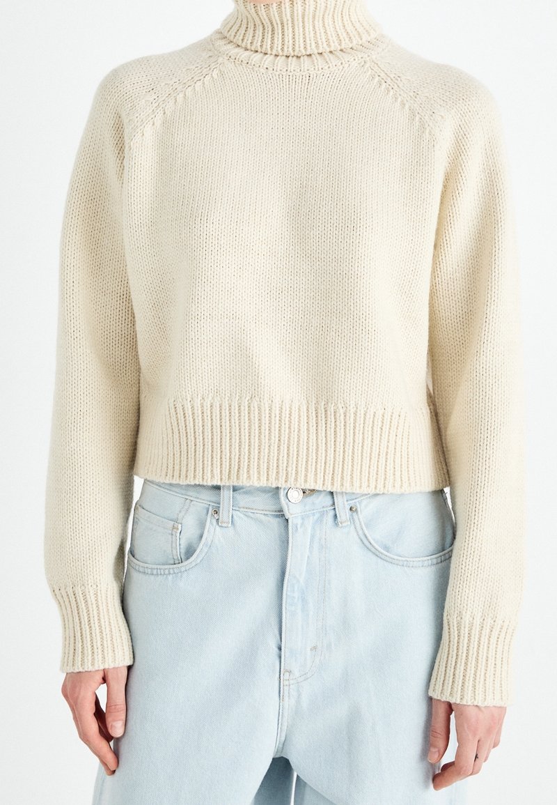 Pull à col roulé en grosse maille couleur crème, manches longues, porté par une personne debout, associé à un jean taille haute bleu clair.