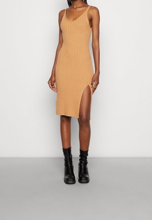 Femme portant une robe midi sans manches côtelée couleur camel avec une fente latérale, associée à des bottes en cuir noires mi-mollet sur fond blanc.