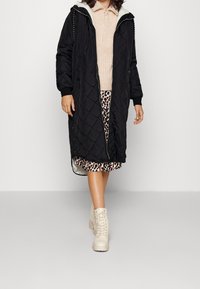 Abrigo largo acolchado negro con capucha, que presenta un cierre de cremallera en la parte delantera y bolsillos laterales. Llevado sobre un top de punto beige y una falda de estampado de leopardo.