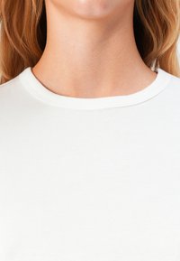 Primo piano di un collo e della parte superiore del torace di una persona con pelle chiara e lunghi capelli biondi, che indossa una maglietta bianca a girocollo.