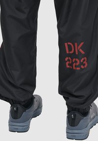 Czarna wodoodporna para spodni o luźnym kroju, z czerwonym nadrukiem "DK 223" po boku. W komplecie z szarymi butami sportowymi o fakturowanej powierzchni.